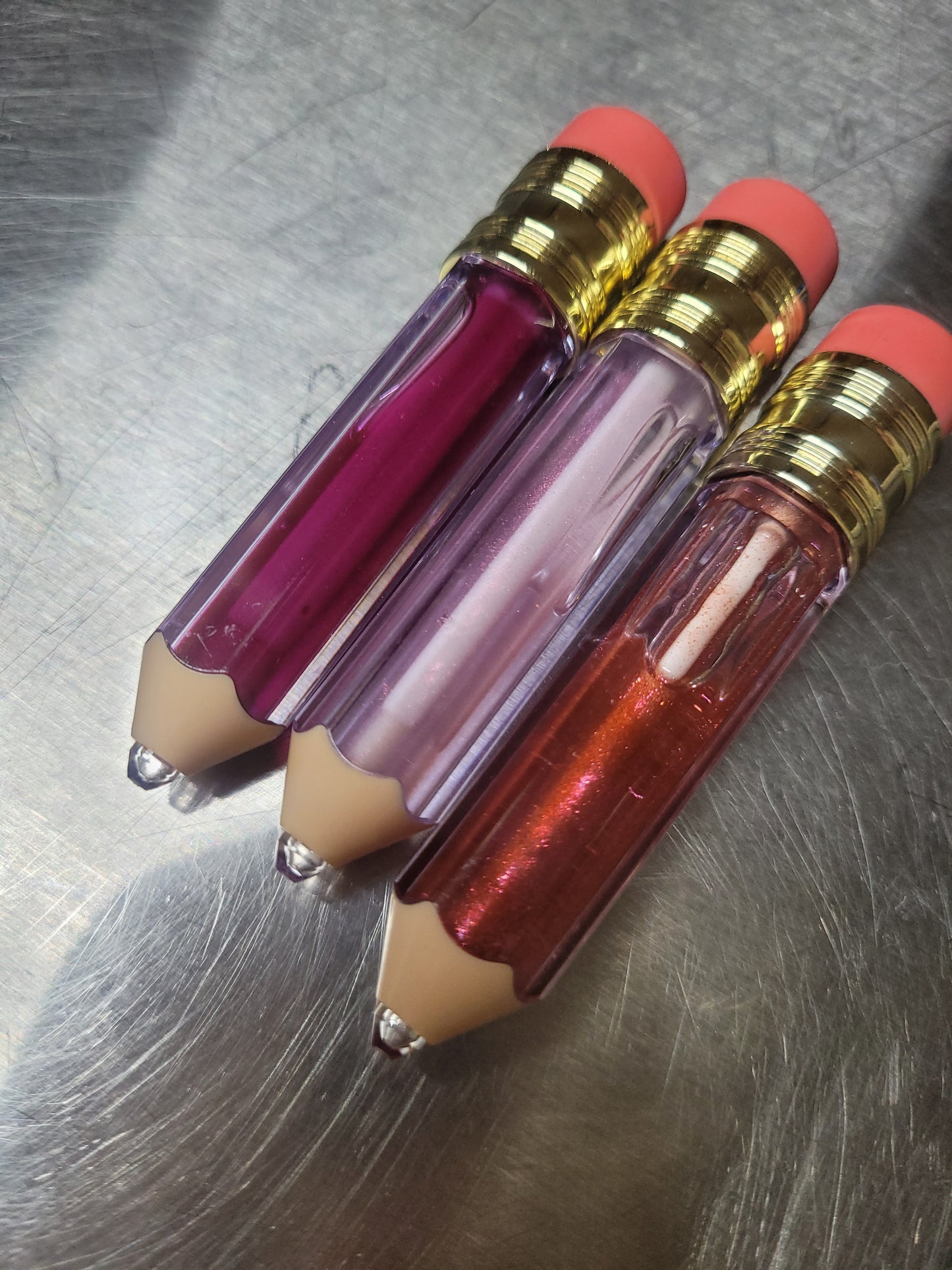 Three lip glosses with pencil-like design on a metallic surface
