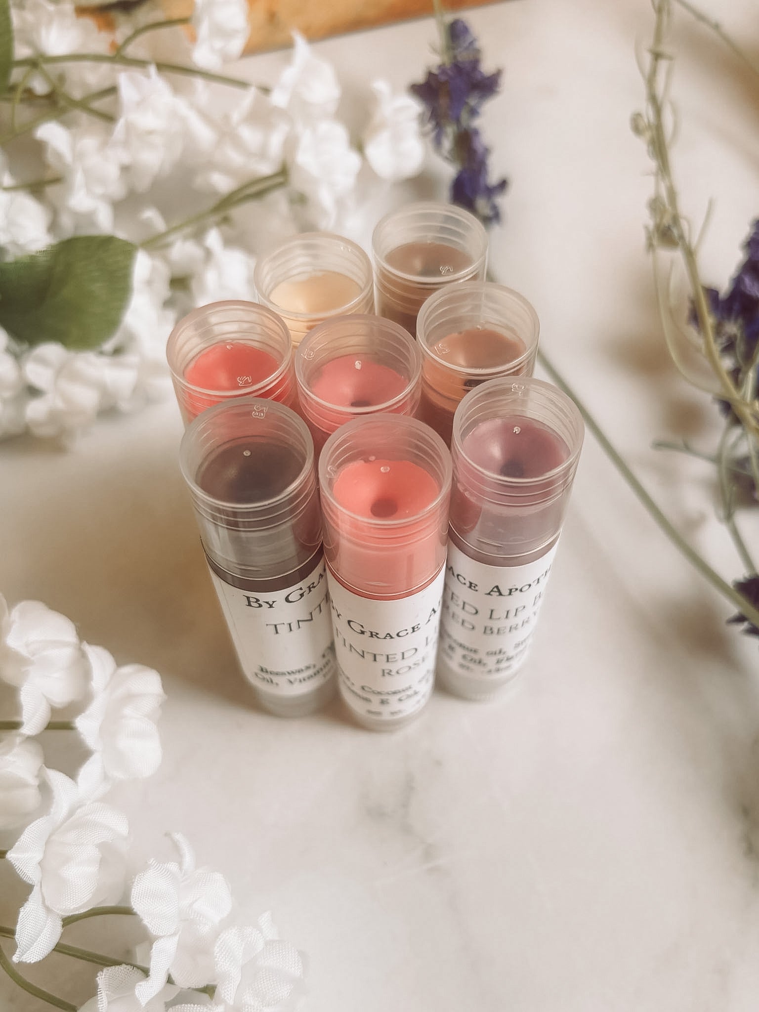Set of lip balm tubes with visible brand names on a marble surface with flowers in the background