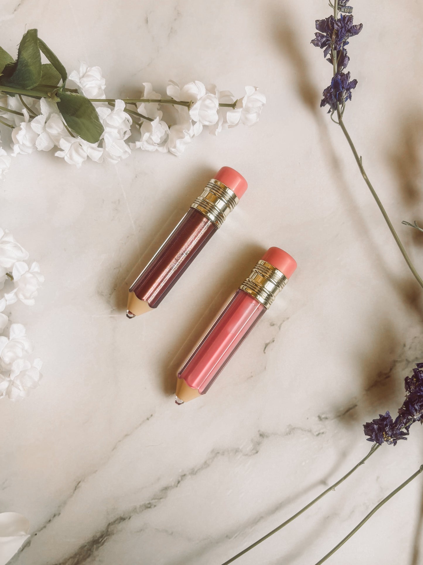Two lip glosses on a marble surface with flowers around