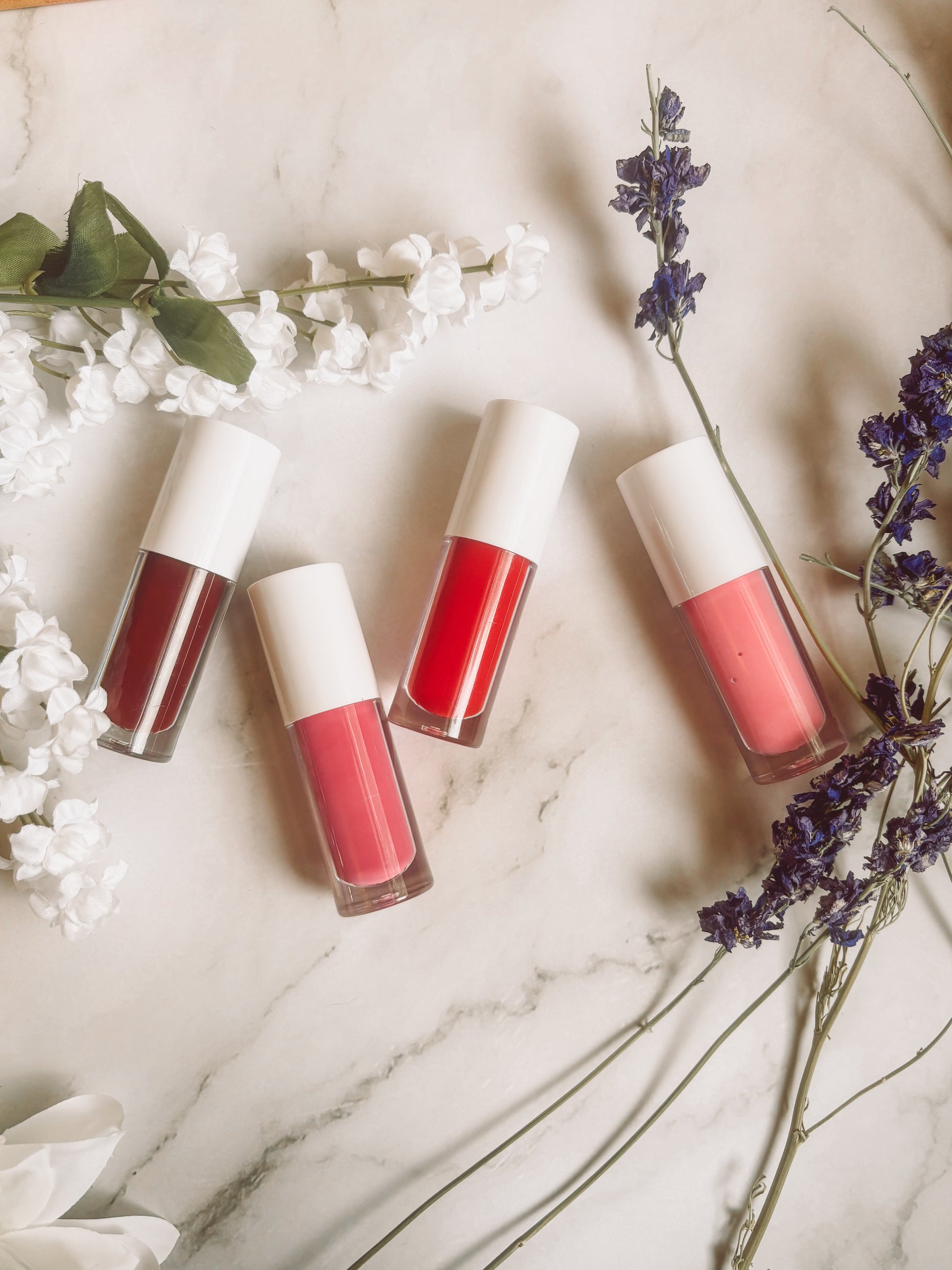 Four lip gloss bottles with different shades on a marble surface with flowers.